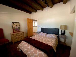 Cozy bedroom in RanchHouse #5 featuring traditional Vigas beams, a comfortable bed with patterned bedding, a dresser with artistic decor, and warm lighting creating a serene ambiance at The Chi Center.