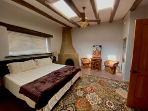 RanchHouse #1 - PORTALES room at The Chi Center featuring a spacious layout with rustic wooden ceiling beams, a traditional adobe-style fireplace, cozy twin beds, southwestern-style chairs, and a vibrant patterned rug for a relaxing retreat.