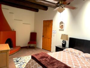 RanchHouse #4 - ADOBE-NICHO room at The Chi Center featuring a cozy bed with patterned bedding, a vibrant red adobe-style fireplace, rustic wooden beams, and a southwestern-style niche with decorative accents.