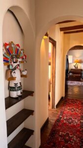 Zia Apartment hallway featuring Southwestern charm. This inviting space highlights a traditional Kachina doll display set against curved adobe walls, with a vibrant, handwoven red rug adding warmth and texture. Archways lead to a cozy, sunlit room adorned with rustic furniture, blending cultural heritage and modern comfort.
