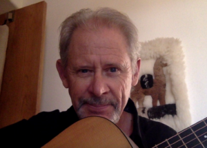 Chris playing a guitar with a warm smile, sitting in a cozy indoor setting with a decorative alpaca-themed wall hanging in the background.