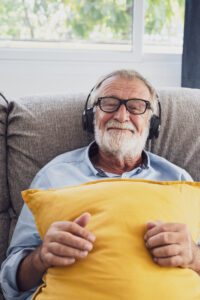 Smiling elderly man with headphones relaxing on a couch, holding a yellow pillow, enjoying a peaceful and uplifting audio experience in a cozy home setting.