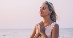 Peaceful woman meditating with hands in prayer position, smiling with eyes closed near the ocean at sunset, symbolizing mindfulness, gratitude, and inner serenity.