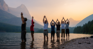 Group of people practicing mindful movements and stretching in a serene lake setting with stunning mountain views at sunrise, symbolizing connection, wellness, and nature-inspired healing.