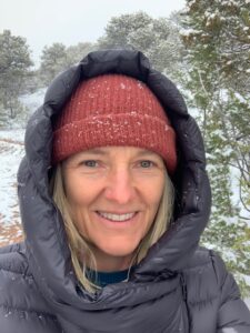 Blyth smiling outdoors in a snowy landscape, wearing a red knit hat and a warm black jacket with a hood, enjoying a winter day.