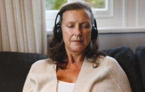 Calm senior woman wearing headphones, sitting on a comfortable couch with her eyes closed, enjoying a peaceful meditation or listening session at home.