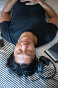 Man lying down on a striped pillow with closed eyes, wearing a black shirt, with headphones and a tablet nearby, enjoying a moment of relaxation or meditation.