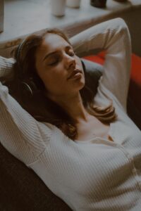 Young woman relaxing with headphones, leaning back on a chair with her eyes closed, enjoying soothing music or meditation in a cozy indoor setting.