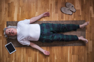 Senior man in plaid pajama pants lying in Savasana pose on a yoga mat at home, with slippers and a tablet nearby on a wooden floor, practicing relaxation or meditation.