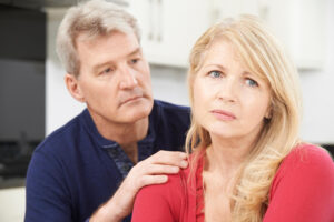 A close-up image of a mature man and woman in an indoor setting, presumably a kitchen. The man, with short gray hair, is wearing a dark blue shirt is looking at the woman. The woman, with long blonde hair, is wearing a red top and looking directly at the camera with a thoughtful expression. The man's hand is gently placed on the woman's shoulder. Both individuals exude a sense of seriousness or contemplation.
