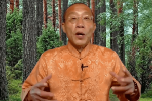 Master Mingtong Gu speaking against a serene forest backdrop, wearing a traditional orange garment, symbolizing wisdom and connection with nature.