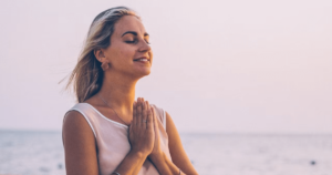A woman with blonde hair is seen in profile against a backdrop of the ocean during sunset. She has her eyes closed and hands clasped in a prayer or meditative gesture. She wears a light-colored sleeveless top, and the warm lighting gives the scene a serene and peaceful atmosphere.