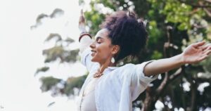 A woman, with a radiant smile and curly hair, gracefully extends her arms amidst a backdrop of lush trees.