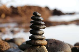 A carefully balanced stack of smooth, dark stones stands in the foreground on a rocky beach. The stack gradually tapers with the smallest stone at the top. The background is blurred, showcasing the shimmering water and rugged coastline, bathed in soft golden light. The overall scene evokes a sense of tranquility and natural beauty.