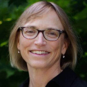 Portrait of a smiling woman with short blonde hair, wearing glasses, earrings, and a black shirt, set against a lush green outdoor background.