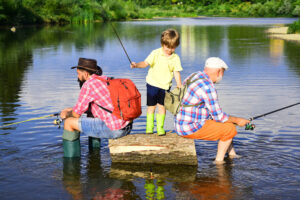 Three generations fishing together by a calm lake, featuring a young boy, his father, and grandfather enjoying quality time outdoors in a serene natural setting.