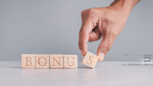 Close-up of a hand placing a wooden block with the letter 'S' to complete the word 'BONUS,' formed with wooden blocks on a reflective surface against a gray background.