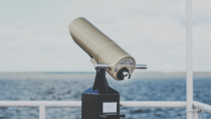 Close-up of a gold public telescope mounted on a black stand, overlooking a vast body of water under a cloudy sky.