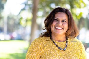 Portrait of a smiling woman with shoulder-length brown hair wearing a yellow patterned blouse and a beaded necklace, set against a blurred outdoor background with trees and sunlight.