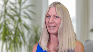 Portrait of Debra Chamberlin Taylor smiling with long blonde hair wearing a blue top, set against a softly blurred indoor background with green plants.