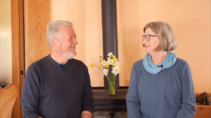 A cheerful older couple sitting indoors, smiling at each other in front of a fireplace with a vase of yellow and white flowers.