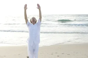 Man practicing Qigong on a sandy beach, dressed in white, with arms raised toward the sky and waves gently rolling in the background.