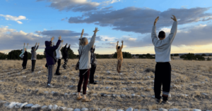 Group of people practicing outdoor Qigong movements in a serene field at sunset, surrounded by nature and stones forming a circular pathway.