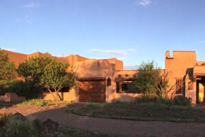 Adobe-style home bathed in warm sunlight, surrounded by greenery and set against a clear blue sky, showcasing traditional Southwestern architecture and serene landscaping.