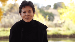 Portrait of a woman in a black sweater speaking outdoors with a serene background of autumn foliage and soft sunlight.