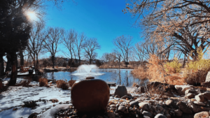 Peaceful winter scene featuring a fountain in the center of a serene pond, surrounded by bare trees, rocks, and snow-covered ground under a clear blue sky