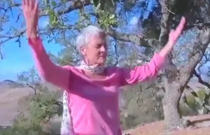 Older woman practicing Qigong outdoors in a peaceful natural setting, surrounded by trees and a clear blue sky.