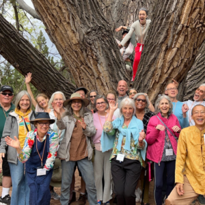 A joyful group of people gathered in front of a large, ancient tree, smiling and waving, celebrating connection and community in an outdoor setting.