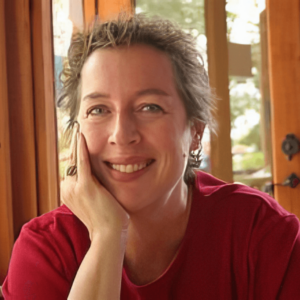 Smiling portrait of Ann Marie, resting her head on her hand, wearing a red shirt, with natural light streaming through wooden-framed windows in the background.