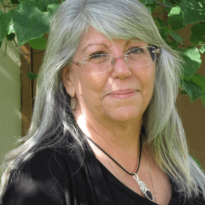 Portrait of Susan Zumo standing outdoors, with silver hair, glasses, and a kind expression, wearing a black top and a silver pendant necklace, set against a natural leafy background.