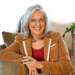 Portrait of Amara Charles smiling warmly, seated on a comfortable chair, wearing a brown blazer and red top, with silver hair and turquoise jewelry, exuding a welcoming and professional demeanor.