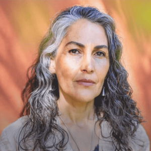 Portrait of a woman with flowing gray and black curly hair, wearing neutral-toned attire, gazing confidently with a warm, sunlit background.