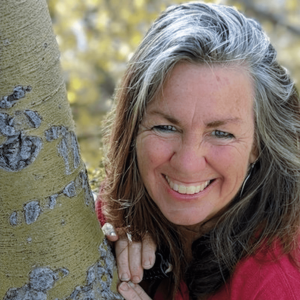 Portrait of a smiling woman with gray hair leaning against a tree, exuding warmth and joy in a natural outdoor setting.