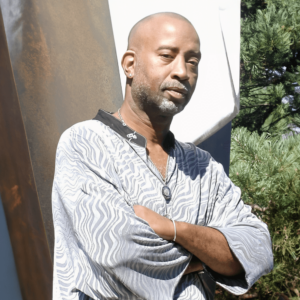 Portrait of a confident man with crossed arms, wearing a patterned shirt and a necklace, standing outdoors against a modern art sculpture and lush greenery.