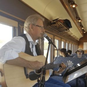 Musician playing an acoustic guitar and singing into a microphone inside a vintage train car, with passengers seated in the background enjoying the performance.
