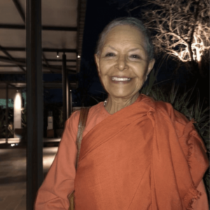 Smiling woman with short hair wearing a red top, standing outdoors at night with subtle lighting and tree silhouettes in the background.