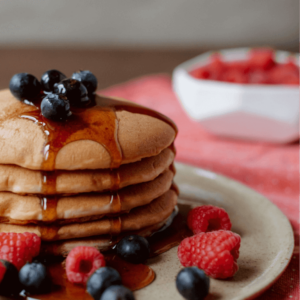 Stack of fluffy pancakes drizzled with maple syrup, topped with fresh blueberries and raspberries, served on a rustic plate with a bowl of fruit in the background.