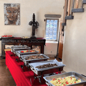 Buffet table with an assortment of breakfast dishes, including scrambled eggs, sausages, and fresh fruit, set against a serene interior featuring a Buddha artwork and rustic decor.
