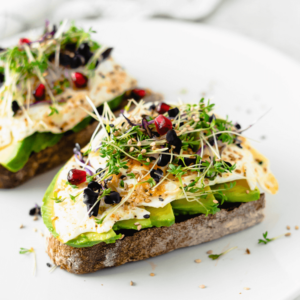 Avocado toast topped with fried eggs, fresh microgreens, sesame seeds, and vibrant pomegranate arils on rustic whole-grain bread for a nutritious and colorful meal.