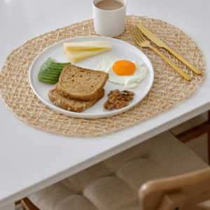 Healthy breakfast plate featuring a sunny-side-up egg, avocado slices, whole-grain toast, walnuts, and cheese sticks, paired with a cup of coffee on a woven placemat and white table setting.