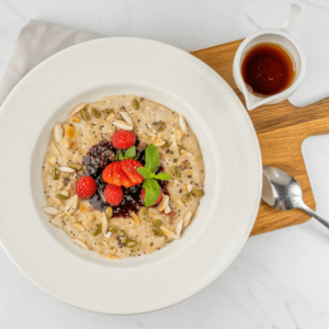 Nutritious oatmeal bowl topped with fresh strawberries, raspberries, mint, almond slices, pumpkin seeds, and a dollop of jam, served with a side of maple syrup on a wooden board.