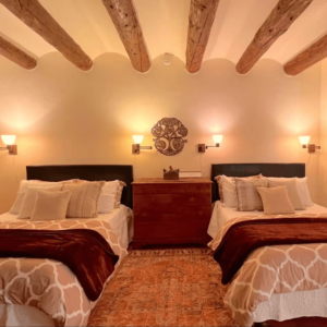 Cozy bedroom with two double beds adorned with soft beige and white bedding, warm lighting, wooden beams, and a decorative tree of life wall art creating a serene retreat ambiance.