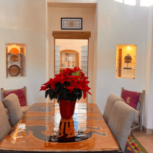 Elegant dining area featuring a polished wooden table with a vibrant poinsettia centerpiece, cushioned chairs, and artistic wall niches displaying decorative plates and a Buddha sculpture, bathed in natural light.