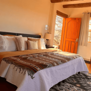 Sunlit guest room with a cozy bed featuring layered pillows and a Southwestern-patterned blanket, complemented by rustic wooden beams and a warm, inviting open door leading to serene outdoor views.