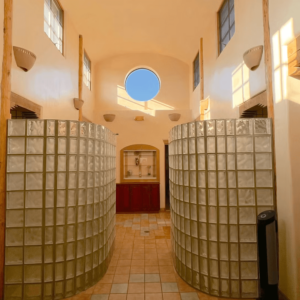 Bright and airy hallway featuring elegant curved glass block partitions, high ceilings with natural wood beams, a circular window inviting sunlight, and rustic tile flooring leading to a decorative alcove.
