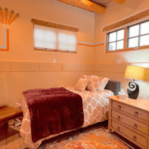 Cozy single-bedroom featuring a comfortable bed with patterned bedding and a plush burgundy throw, complemented by natural wood beams, soft lighting from a modern lamp, and rustic Southwestern decor.